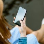 A young woman holding a smartphone while sitting outdoors in summer, capturing a moment of relaxation and technology use.