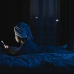 A woman lying in bed with a towel on her head, using a mobile phone at night.