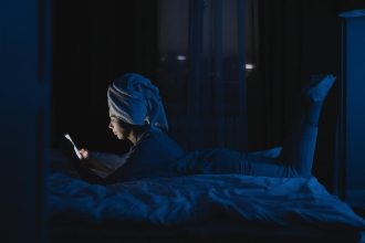 A woman lying in bed with a towel on her head, using a mobile phone at night.