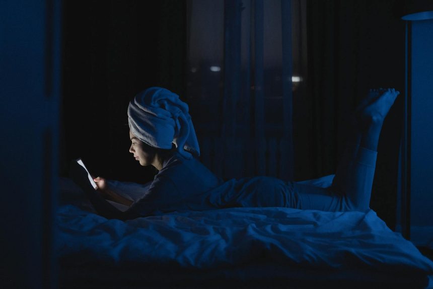 A woman lying in bed with a towel on her head, using a mobile phone at night.