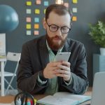 Man in glasses using smartphone at office desk.