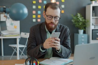 Man in glasses using smartphone at office desk.