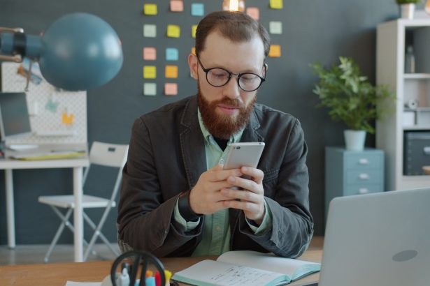 Man in glasses using smartphone at office desk.