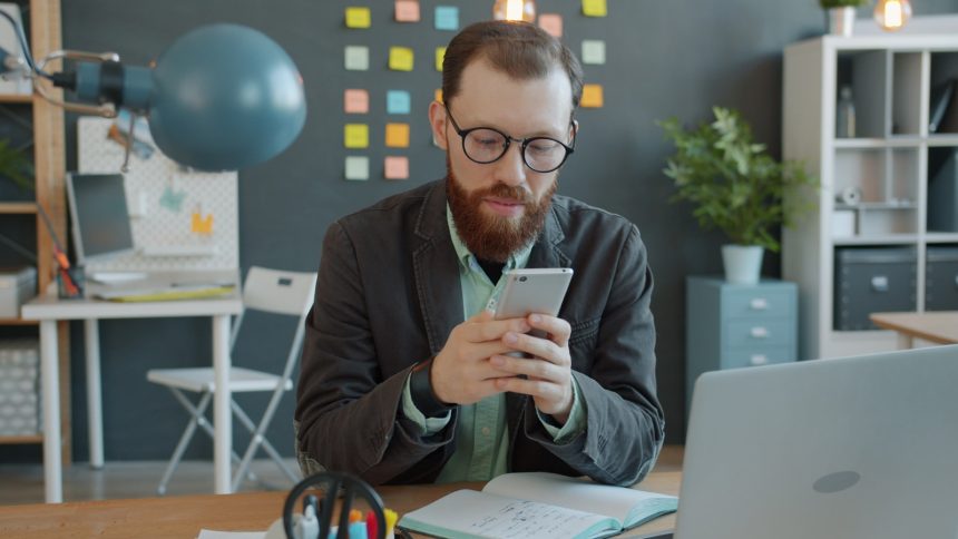 Man in glasses using smartphone at office desk.
