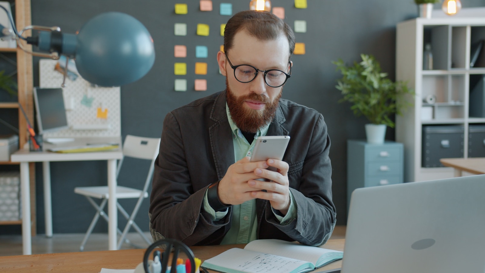 Man in glasses using smartphone at office desk.