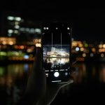 Hand holding phone capturing city lights at night.